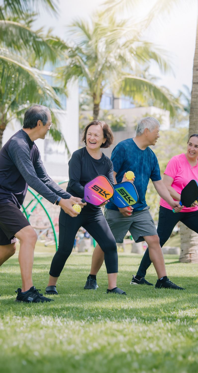 Four pickleball players laughing together holding paddles at a tropical Hawaiʻi court
